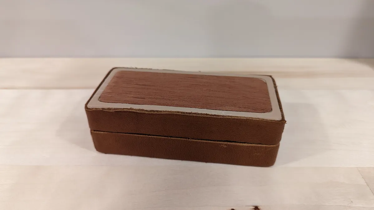 The brown dice box closed, showing its textured lid and rounded rectangular shape.