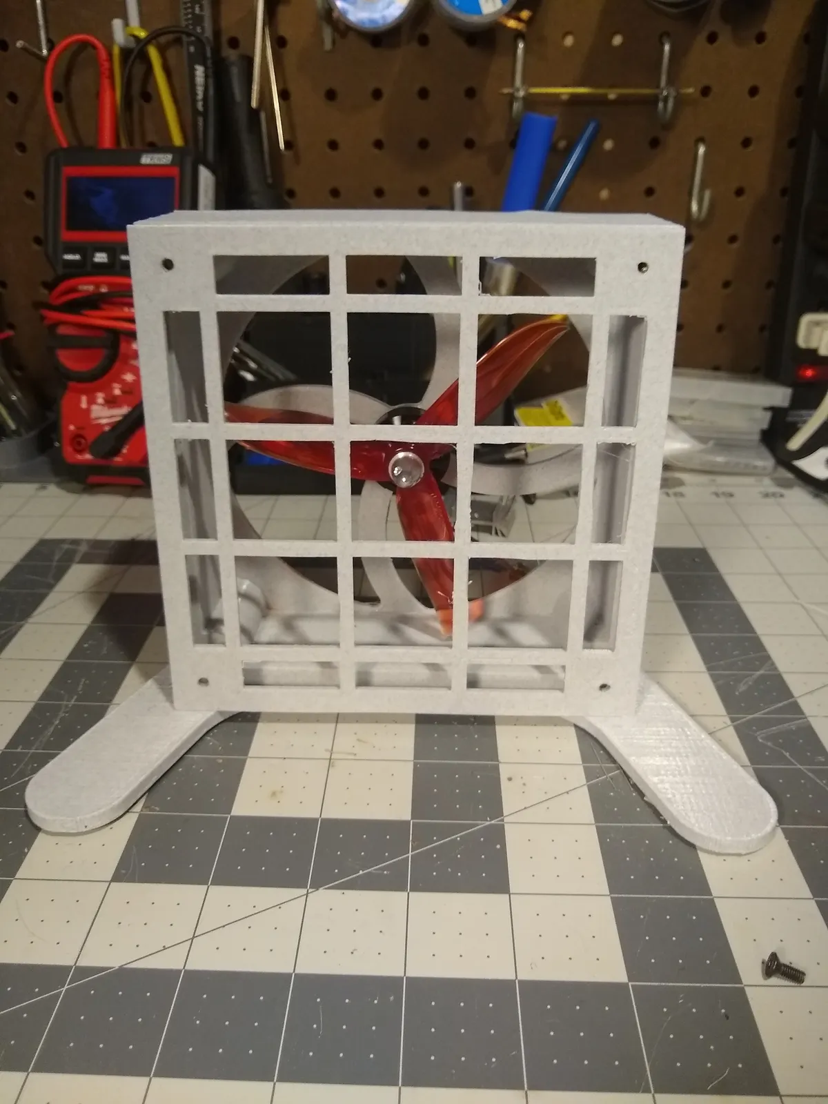 A workshop fan with a white printed housing, front grille, and wide feet on a cutting mat.
