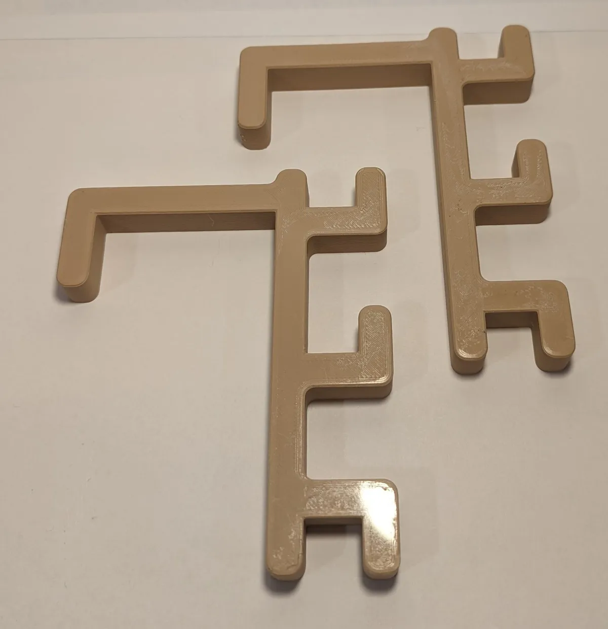 Two tan printed holder arms with long top hooks for hanging over a loom frame and stepped hooks down their sides.
