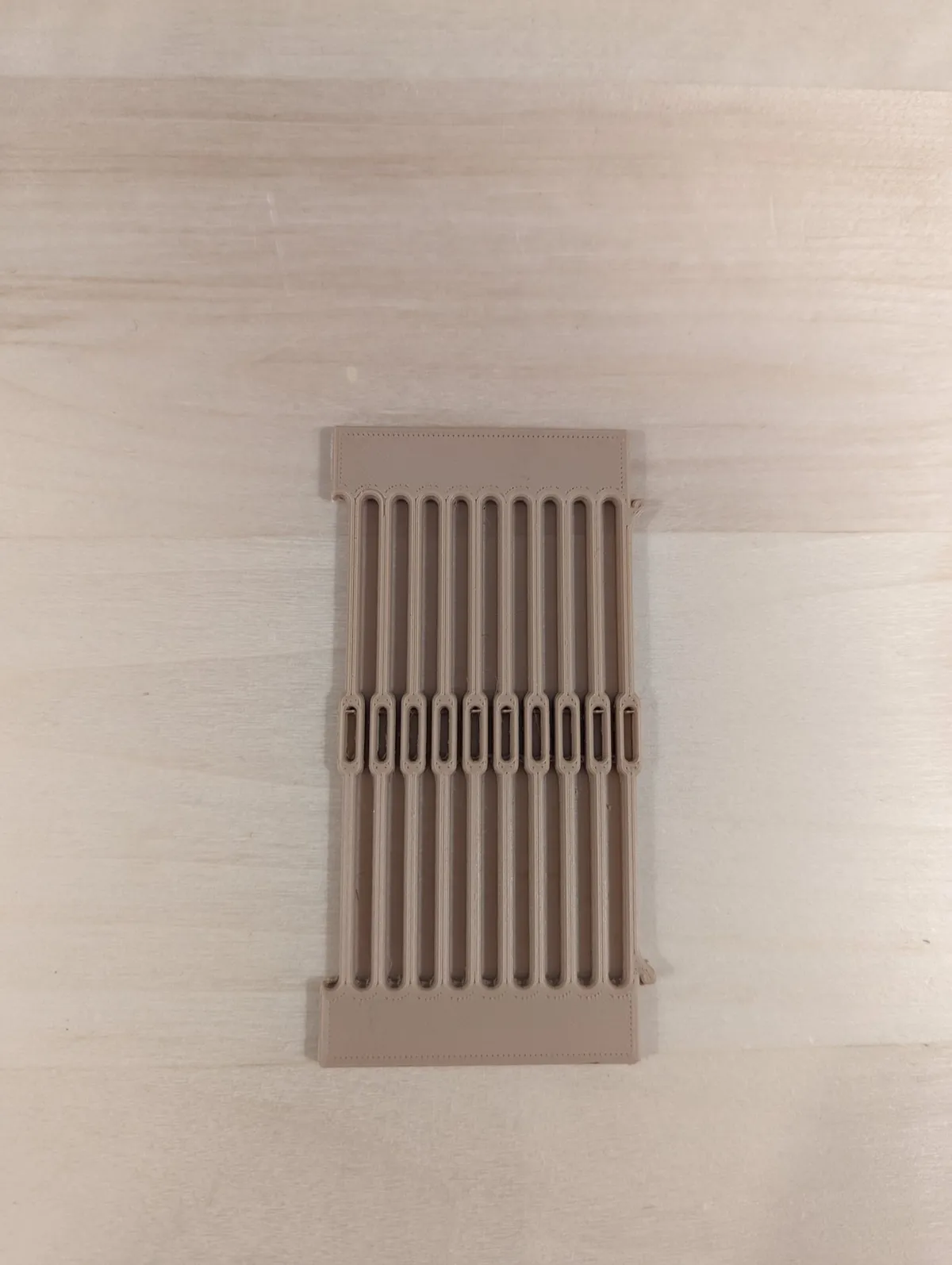 A small narrow tan printed reed with nine slots and eyelets between them, lying on a light wood surface.