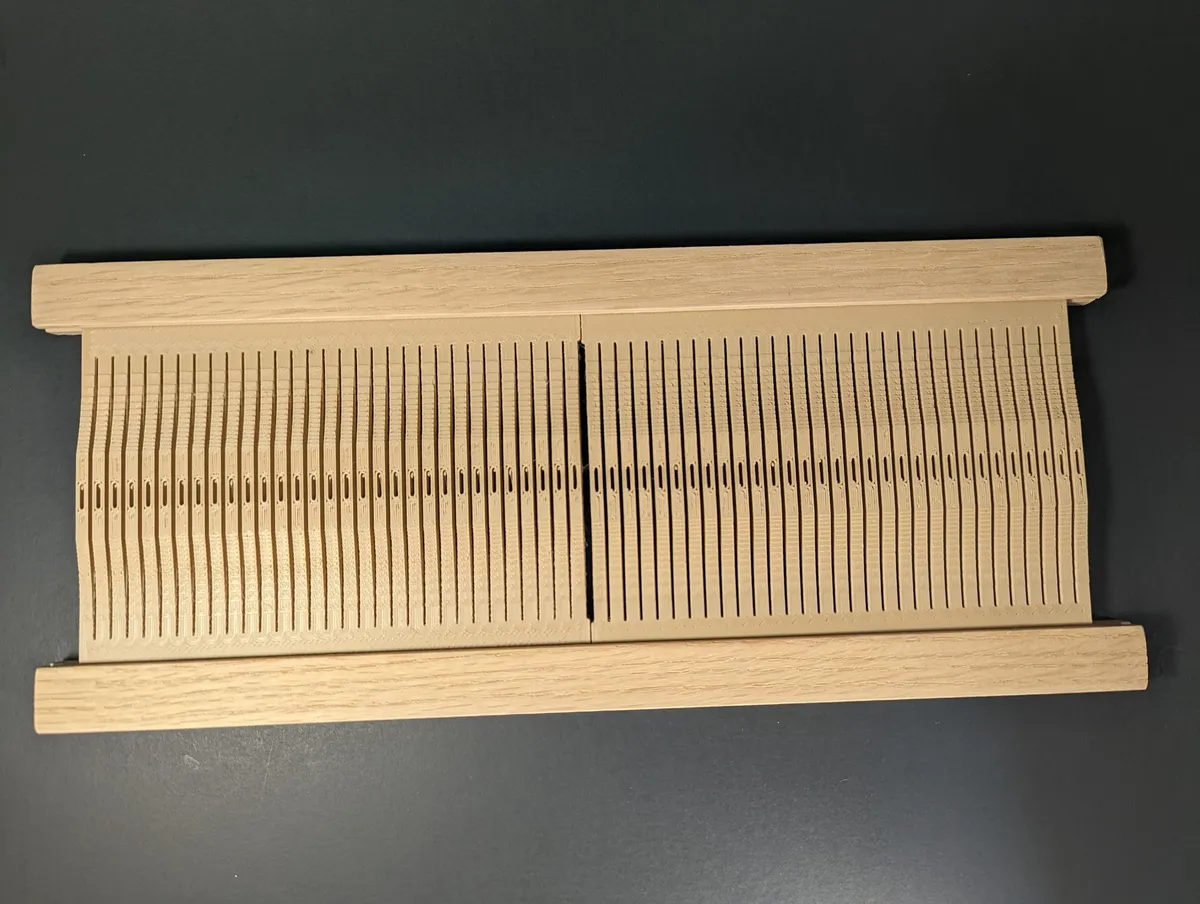 A full-width rigid heddle made from several printed slotted modules held between two light wooden rails on a dark surface.