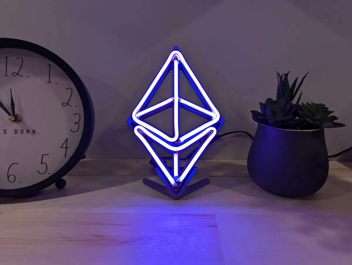 A glowing blue and white Ethereum-shaped desk light in a printed frame beside a clock and plant.