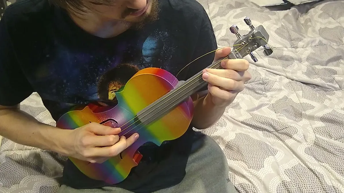 Playing a rainbow-colored printed violin fitted with a gray neck, four strings, and guitar tuning machines.