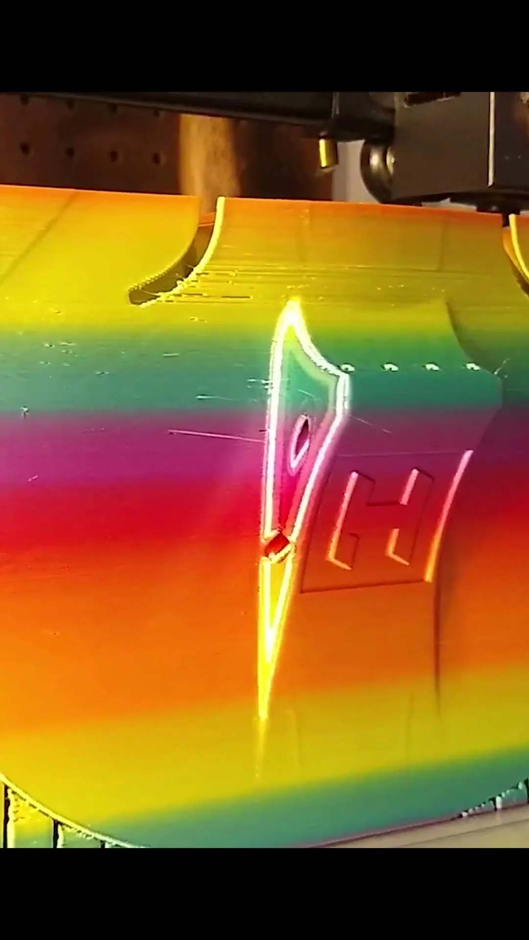 A 3D printer working on the upright rainbow violin body, with its sound holes and raised tailpiece visible.