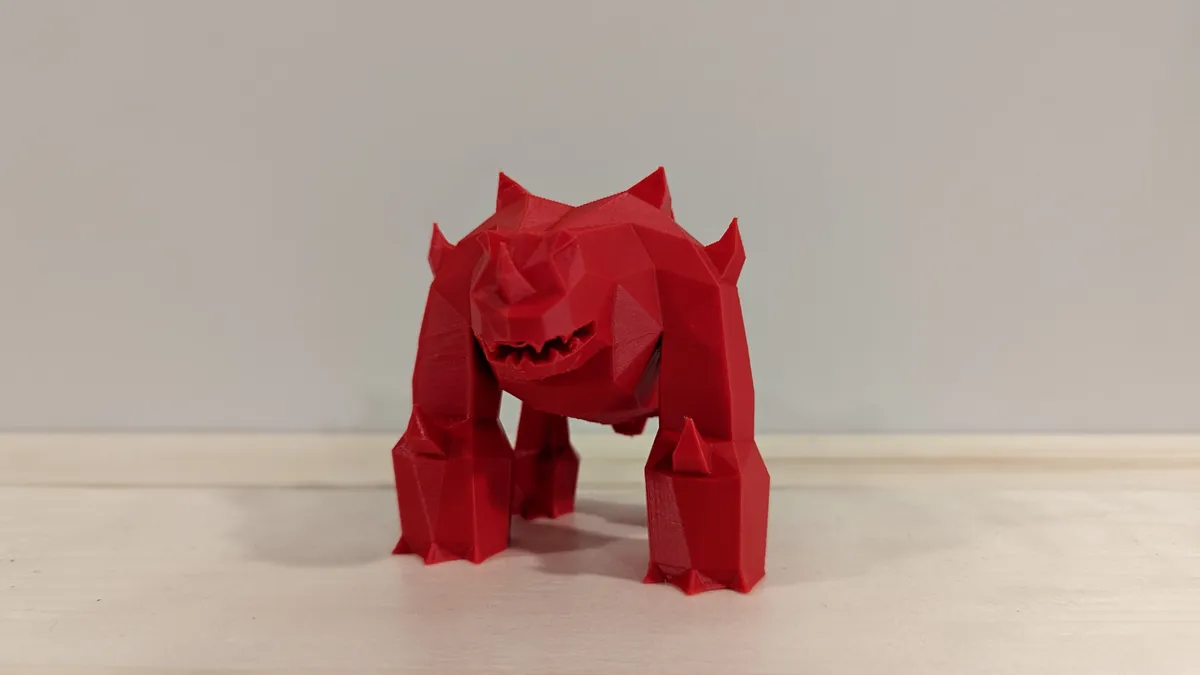 A red 3D-printed Jad figure with angular legs, horns, and an open mouth on a wooden surface.